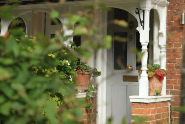 A modern Victorian front door porch is seen through the soft focus evergreen trees in the foreground. Terracotta pots filled with trailing wild strawberries sit on either side of the french grey painted