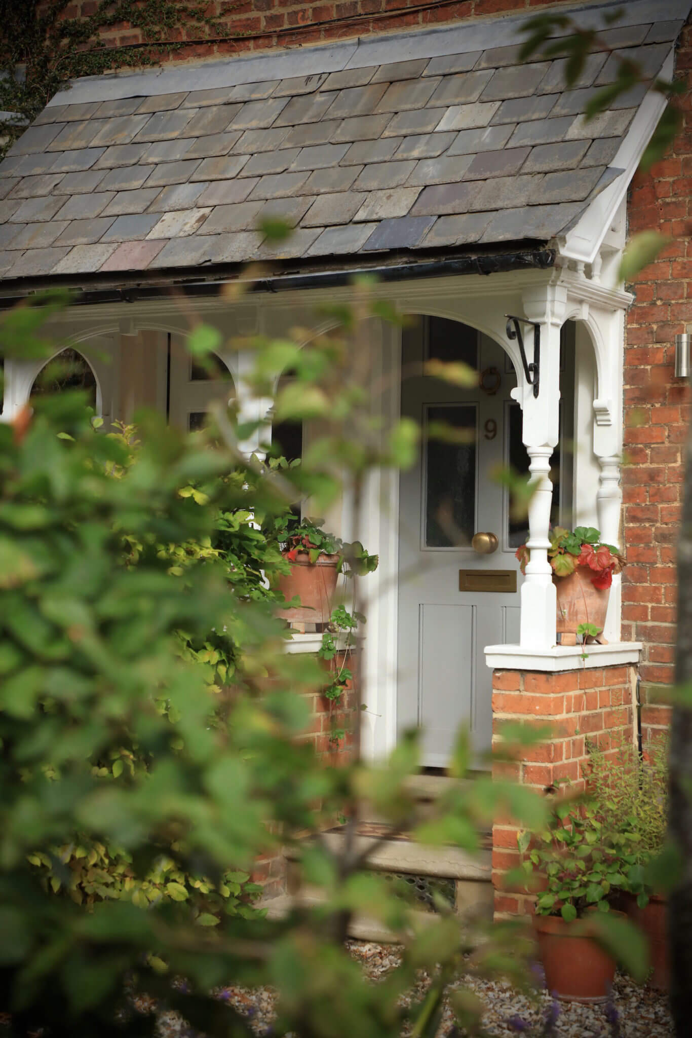 A modern Victorian front door porch is seen through the soft focus evergreen trees in the foreground. Terracotta pots filled with trailing wild strawberries sit on either side of the french grey painted