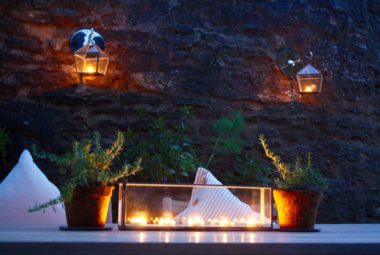 a candle lit courtyard garden with white cushions and lanterns on a stone wall. Tiny Rosemary plants are on the garden dining table