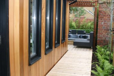 Side passage of wooden cladded building with black framed window. A row of ferns