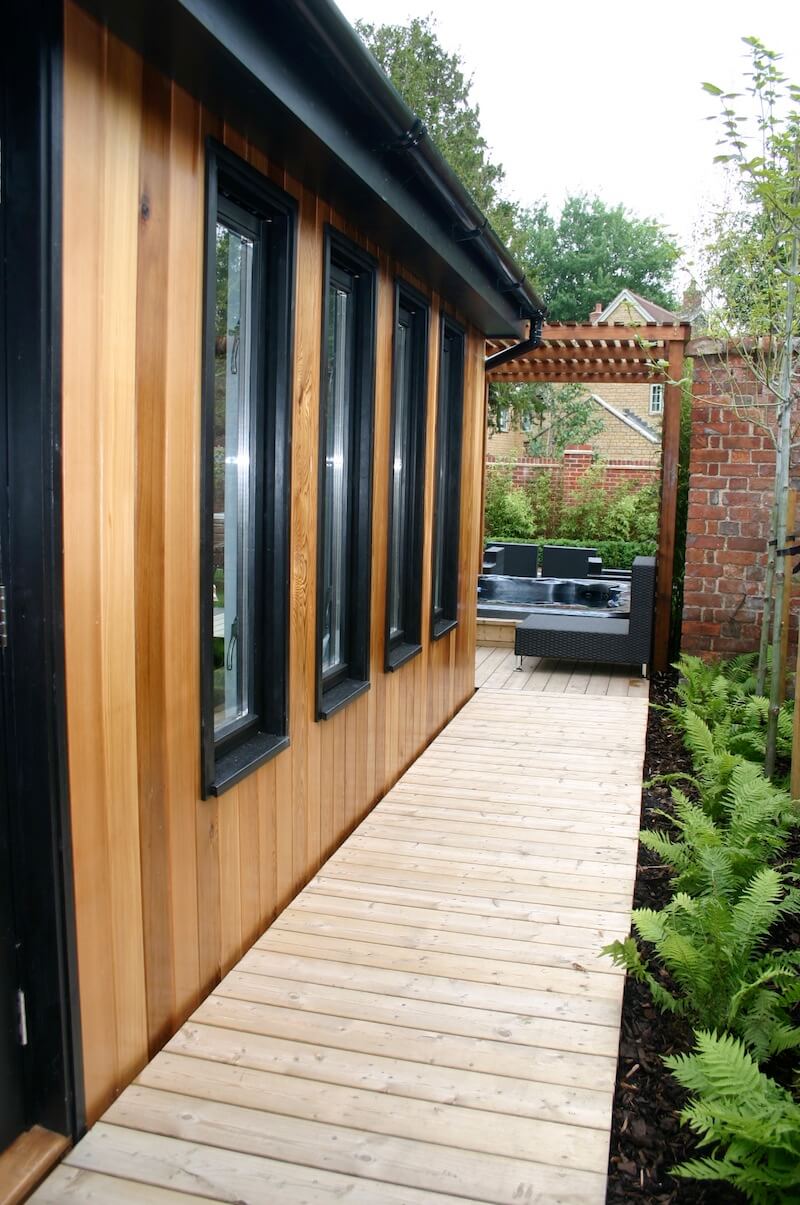 Side passage of wooden cladded building with black framed window. A row of ferns