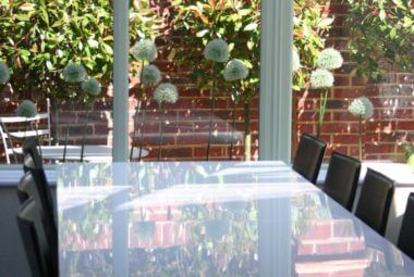 Inside a glass house with garden furniture, through the window are white allium heads