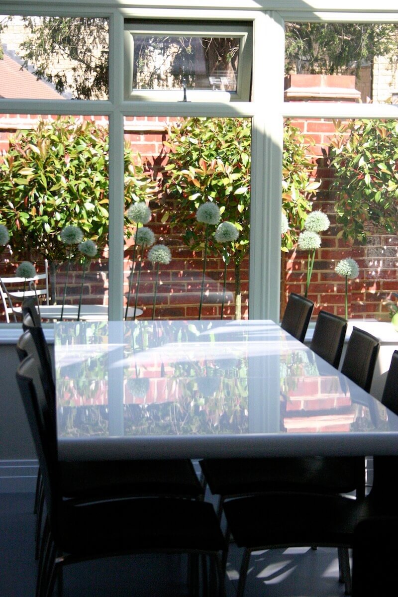 Inside a glass house with garden furniture, through the window are white allium heads