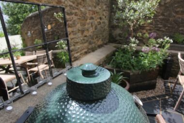 Green barbecue in front of framed garden mirror. In the reflection of the mirror you can see a wooden table and chairs