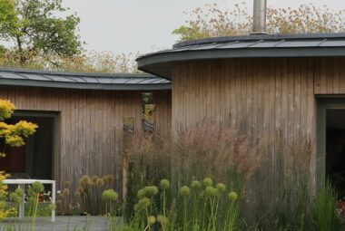 a wood clad house with a living roof and chimney in summer, lawn and slate patio surround it