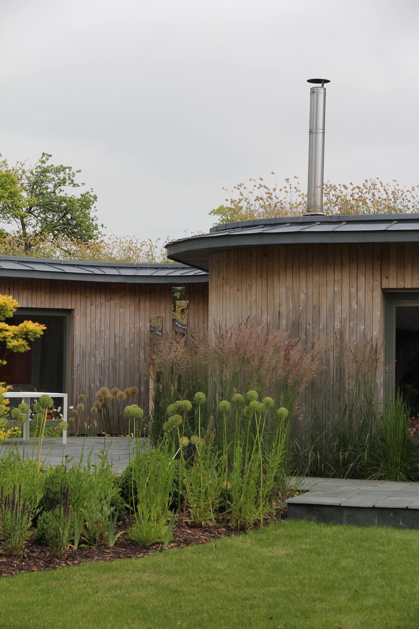 a wood clad house with a living roof and chimney in summer, lawn and slate patio surround it