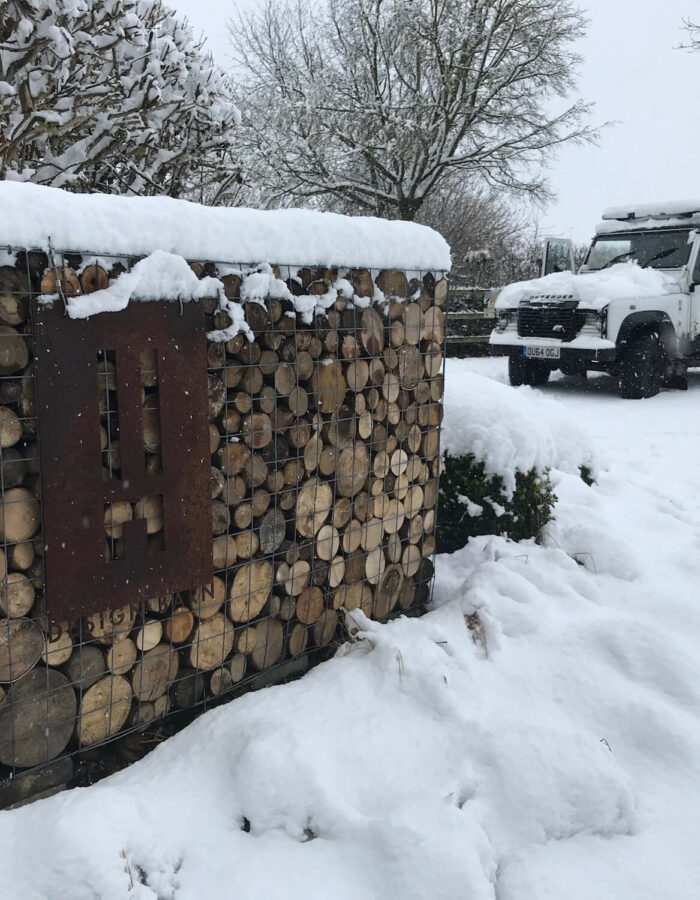 a log gabion with a corten design barn sign and black and white Land Rover Defender all covered in snow in the Cotswolds