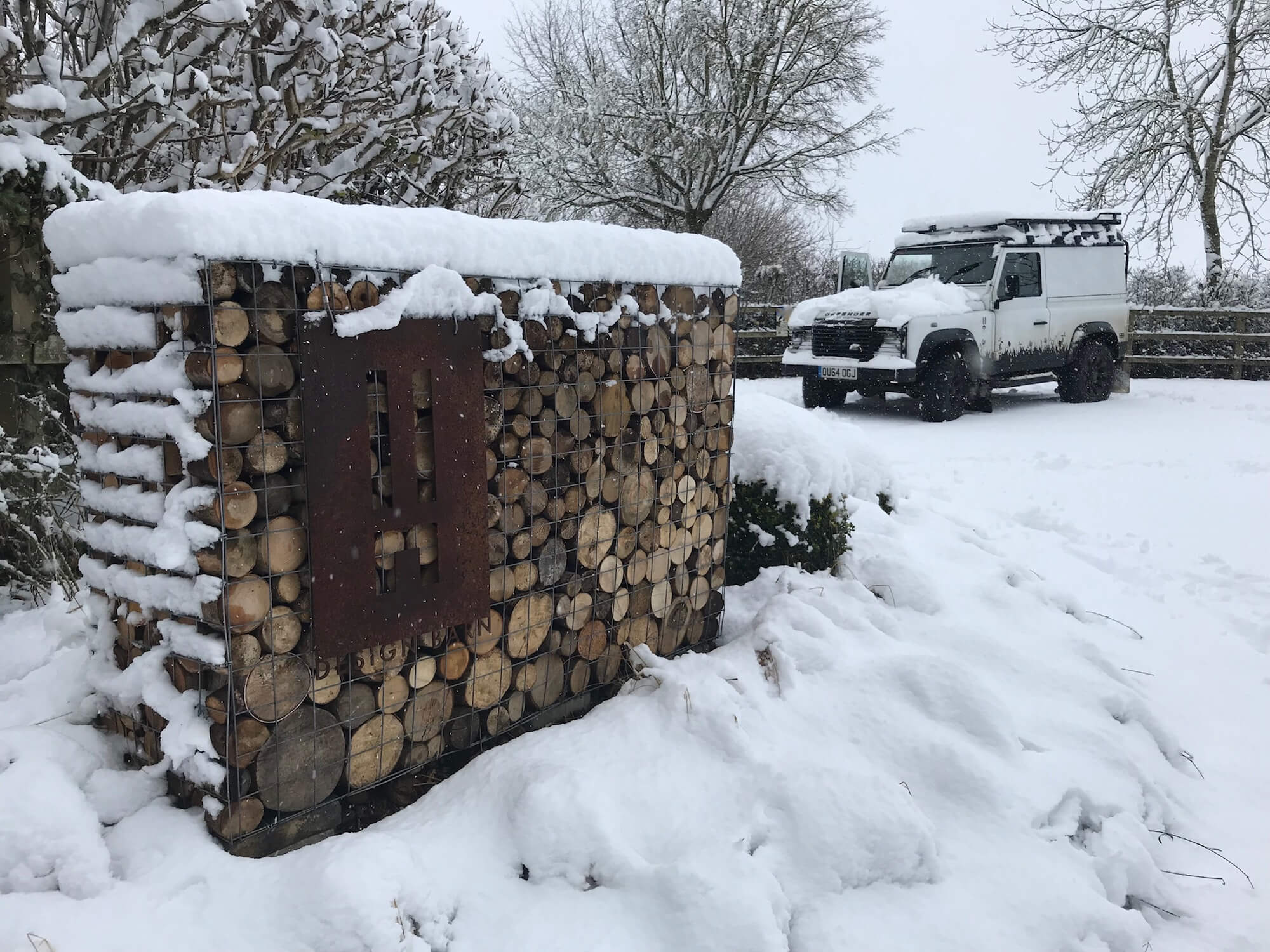 a log gabion with a corten design barn sign and black and white Land Rover Defender all covered in snow in the Cotswolds