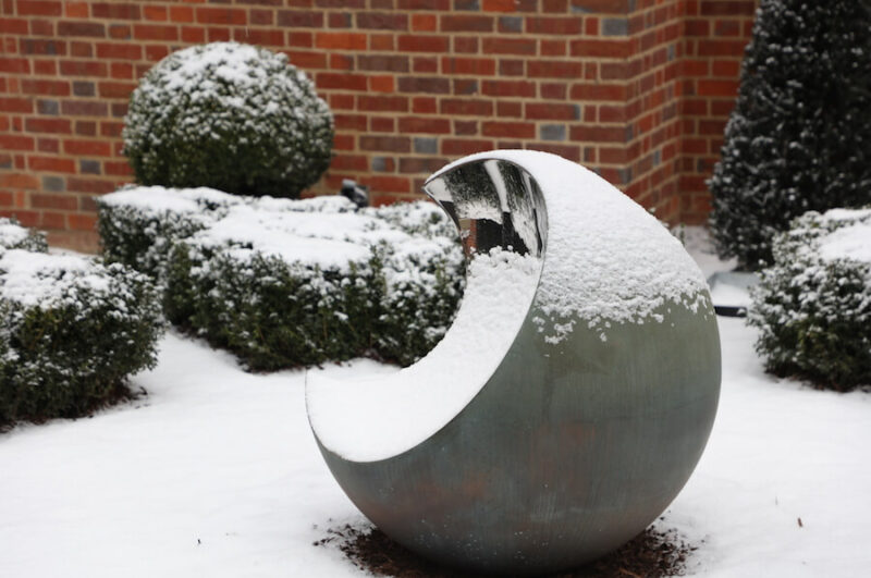 David Harber Bite sculpture covered in snow, a copper and chrome ball garden sculpture in a courtyard