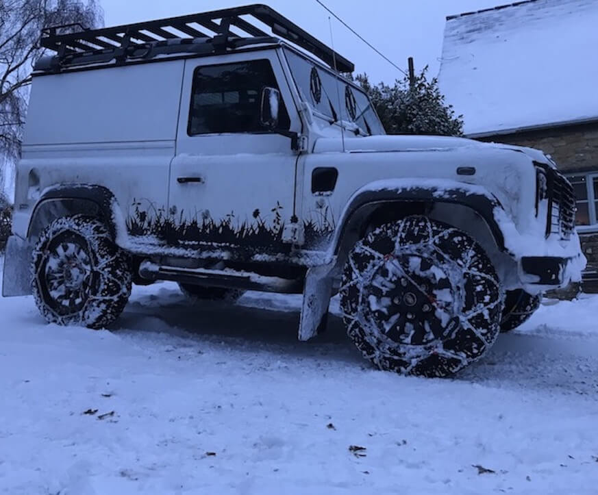 a black and white Land Rover Defender truck with landscape design van graphics in a snowy cotswolds garden with snow chains