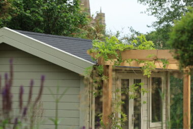 garden building with stone wall and wooden pergola with a wisteria trained. view of church in background